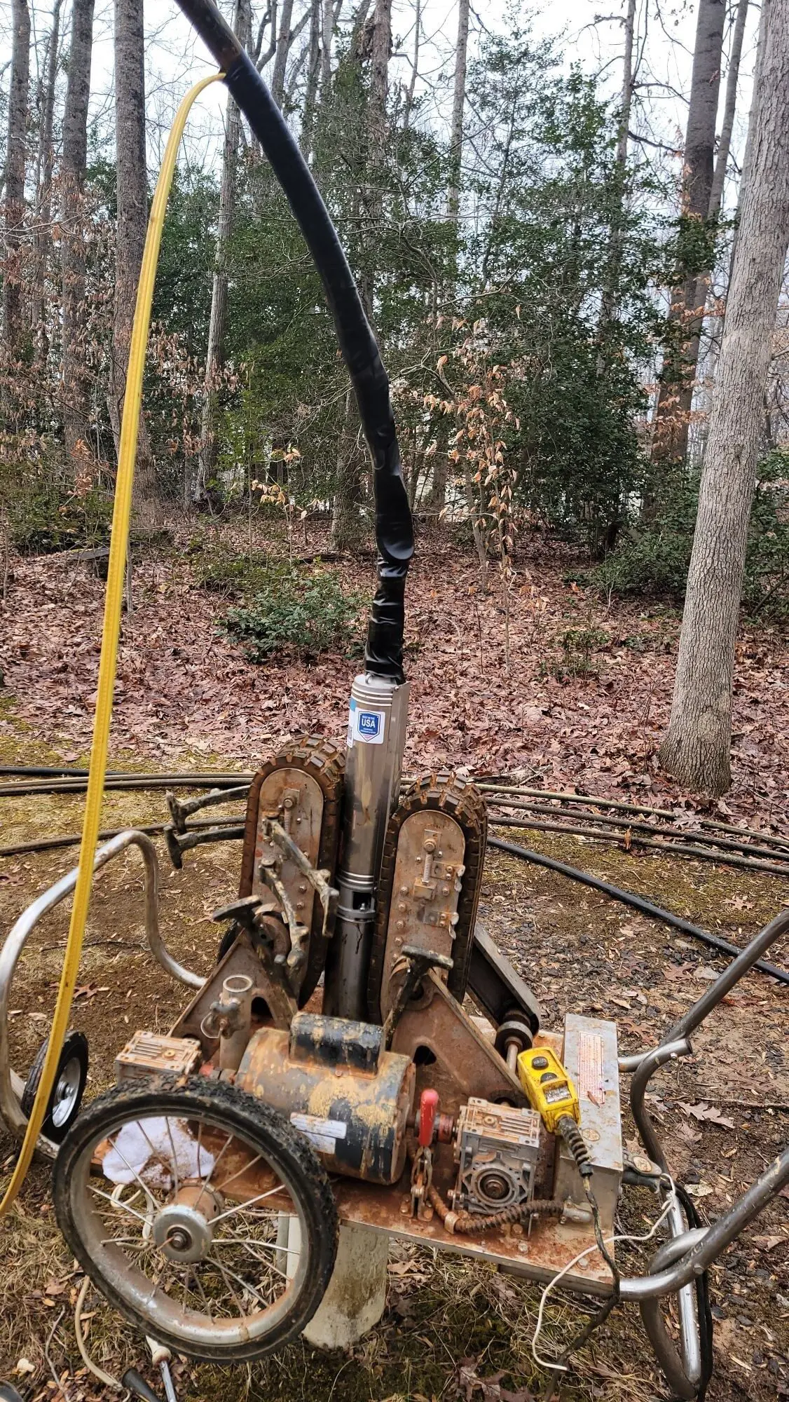 well pump Well-drilling setup in a wooded yard showing a submersible well pump mounted on a drilling rig trolley with hoses and electrical lines connected, surrounded by trees and leaf-covered ground, illustrating residential well pump installation and service