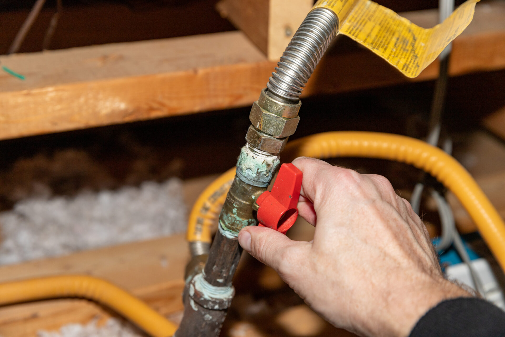 Hand turning gassafetly valve off Hand turning a red shut-off valve on a copper gas/water line in an attic crawlspace, showing plumbing fittings, flexible yellow gas line, and insulation—home plumbing service and repair context.