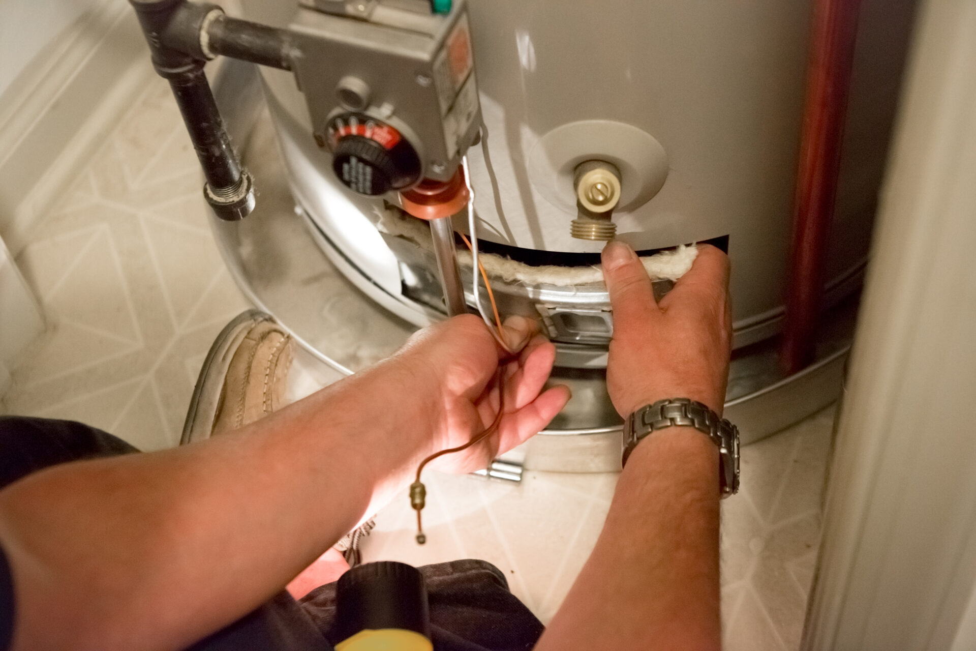 Man shines flashlight on hot water heater Technician repairing a home water heater—hands using tools to adjust wiring and fittings at the tank’s lower access panel in a small utility space