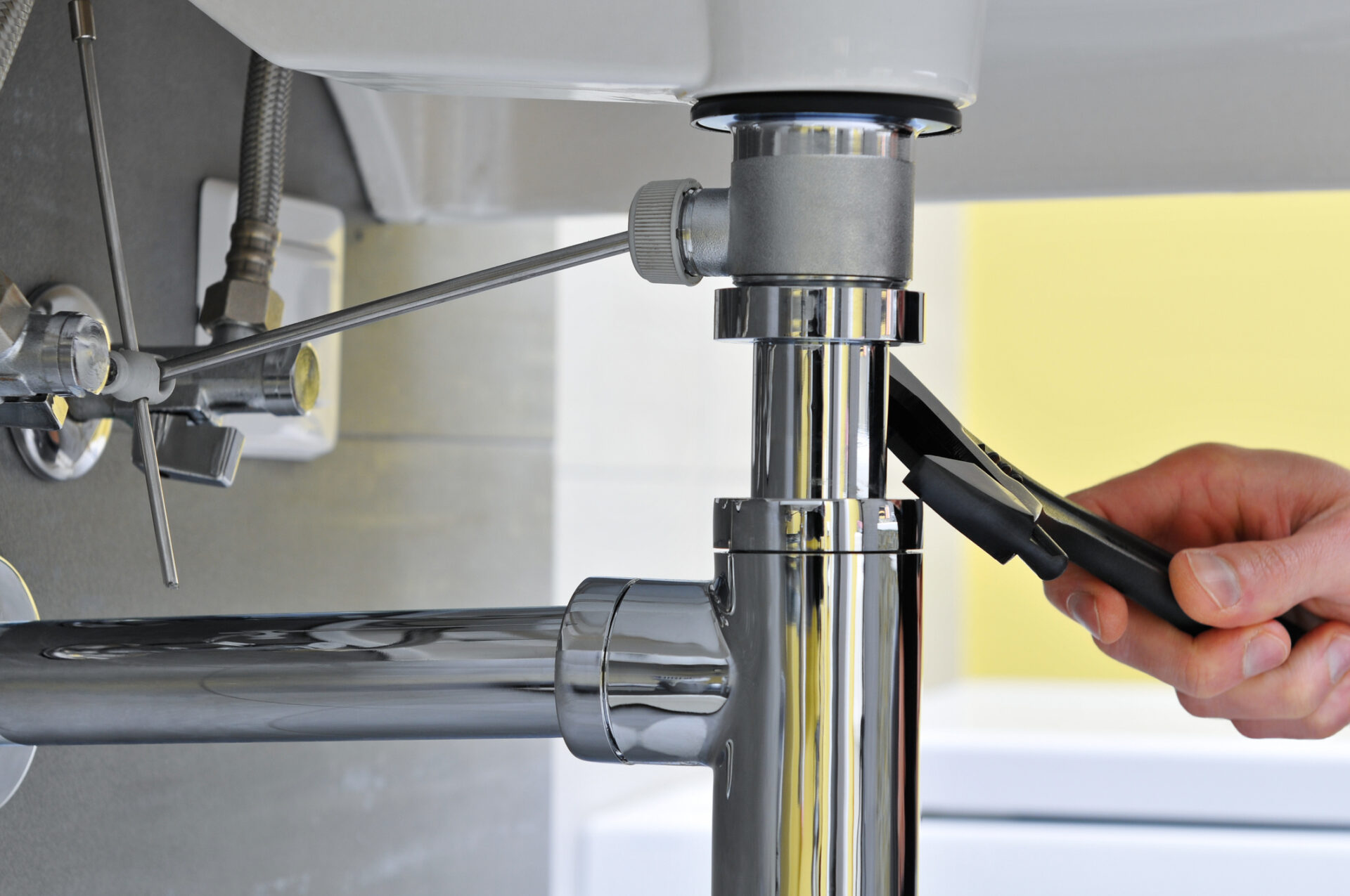 Close-up of plumber's hand doing repairing work at the sink Hand tightening a chrome sink P-trap under a kitchen or bathroom sink with a wrench, showing polished plumbing fixtures, supply lines, and disposal connection — professional faucet, sink, and fixture repair/installation scene